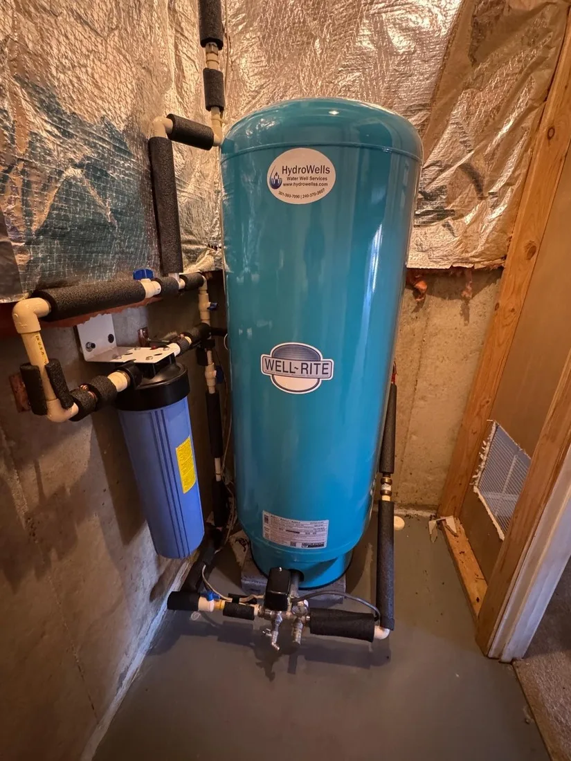 Teal well pressure tank with a HydroWells label installed next to a whole-house sediment filter in a Maryland basement